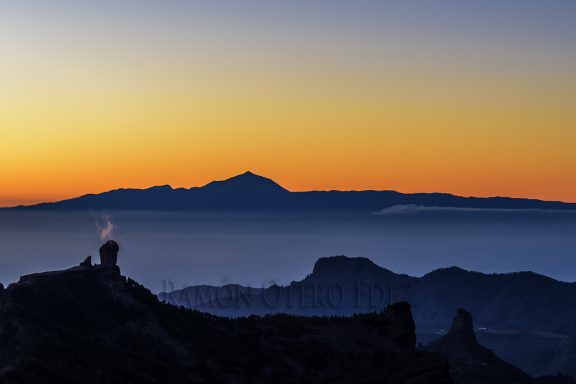 Siluetas de montañas al atardecer con humo saliendo de una cima.